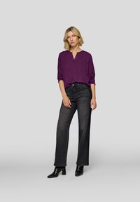 Lila Bluse mit V-Ausschnitt, zwei Fronttaschen und hochgekrempelten Ärmeln; kombiniert mit hochtaillierten dunklen Jeans und schwarzen, knöchelhohen Stiefeln mit Absatz.