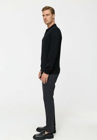 Mustad pikad varrukad ja kaelusega must kudumipolo, mis on kombineeritud hallide kitsaste pükstega ja mustade mokassiinidega, valge taustaga.