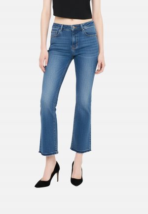 Donna che indossa jeans blu a vita media, taglio svasato e orlo grezzo, abbinati a scarpe nere con tacco alto e punta appuntita, in piedi su sfondo bianco.