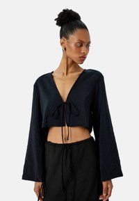 Krótką czarną bluzkę uszytą z teksturowanej tkaniny, z głębokim dekoltem w kształcie litery V, długimi rękawami oraz detalem w postaci związania z przodu w talii.