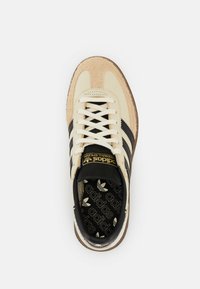 Crèmekleurige suède en textiel sneakers met zwarte strepen en veters, bruine rubberen zool en gouden branding op de tong.