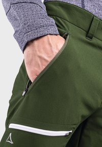 Grüne Outdoor-Hosen mit glatter Textur, ausgestattet mit einer seitlichen Reißverschlusstasche und einem weißen, zippered Akzent. Ein graues Langarmshirt ist teilweise sichtbar.