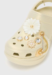 Sabot beige décoré d'une fleur blanche, de perles et de cristaux, avec un pendentif chaîne dorée sur la lanière avant.