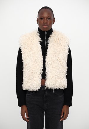 Mouwloze crème nepbont gilet met een gestructureerd oppervlak, gedragen over een zwarte coltrui, gecombineerd met donkerdenim jeans. Ritsluiting bij de hals.