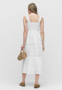 Witte katoenen midi-jurk met broderie anglaise details, brede banden en een smock-borsten. Gecombineerd met een geweven handtas en bruine schoenen.