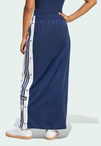 Mörkblå idrottsrock av slätt tyg, med vita tryckknappar och tre ränder längs sidorna, samt en Adidas-logotyp.