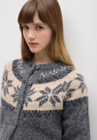 Cardigan lavorato a maglia in grigio con motivo a fiocco di neve in crema e grigio scuro. Presenta una scollatura rotonda e chiusura frontale con bottoni. Tessuto morbido.