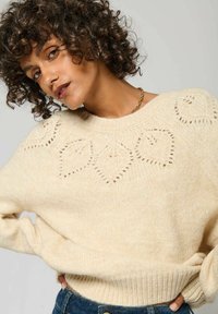 Pull en tricot crème avec des motifs en diamant, poignets et ourlet côtelés. Texture douce, grand col et coupeoversize. Associé à un denim.