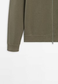 Olijfgroene sweater met ritssluiting en een textuurgebreid ontwerp, voorzien van geribbelde manchetten en zoom, benadrukt een minimalistische stijl.