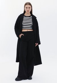 Gehäkeltes Oberteil mit schwarz-weißen Streifen, hoch taillierte schwarze weit geschnittene Hose und ein langer schwarzer Mantel. Das Modell trägt schwarze Plateauschuhe.