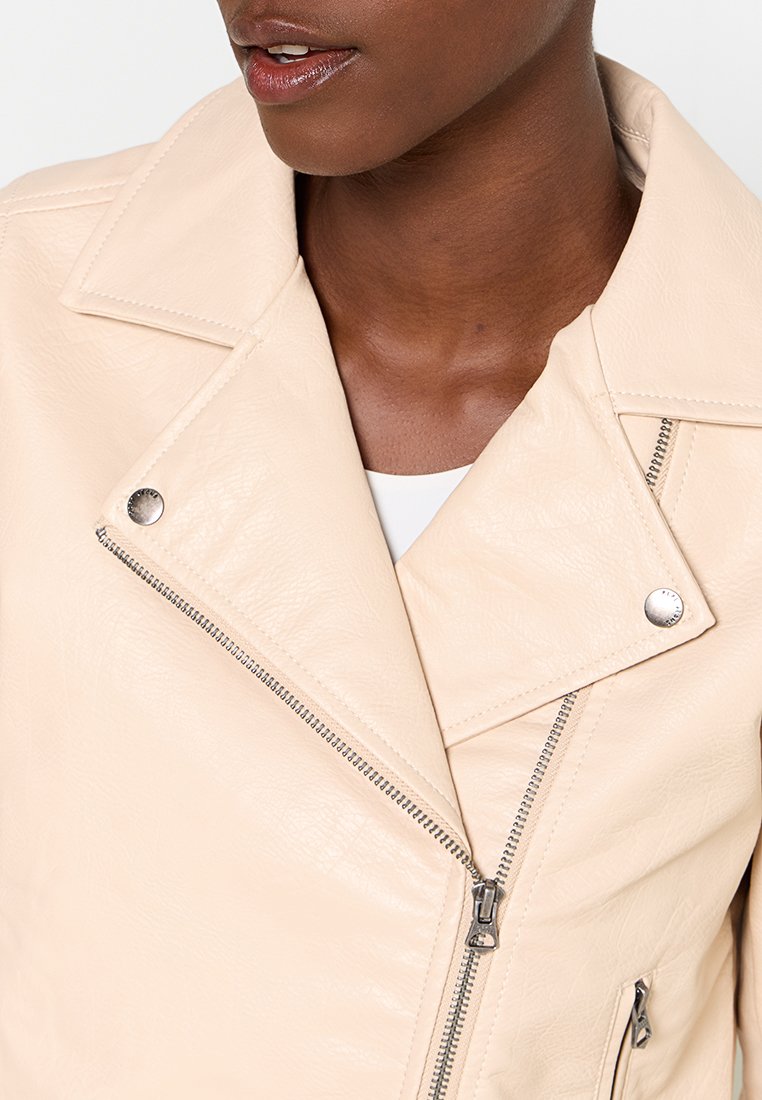 Chaqueta de biker de cuero sintético color crema con herrajes plateados, que presenta un cierre diagonal, solapas de gran tamaño y una superficie texturizada.