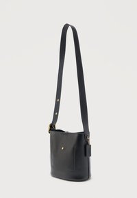 Μαύρη δερμάτινη τσάντα ώμου με ρυθμιζόμενο ιμάντα, μπροστινή τσέπη, χρυσό κουμπί και μικρή κρεμαστή ετικέτα σε λευκό φόντο.