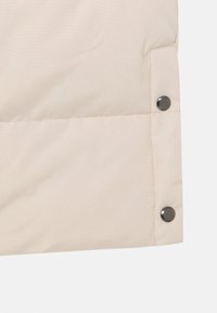 Sección de chaqueta aislante de color beige claro con una textura suave, que presenta botones oscuros y un patrón acolchado horizontal a lo largo de la costura.