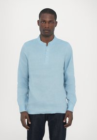 Lyseblå langermet henley-skjorte laget av teksturert stoff, med rund krage og tre-knapps placket. Brukt med mørke jeans.