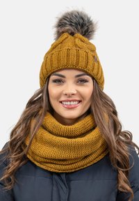 Gorro de punto amarillo mostaza con pompón en la parte superior, combinado con una bufanda infinita texturizada a juego, llevada sobre un abrigo oscuro.