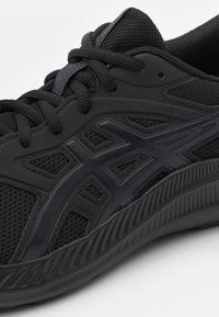 Chaussure de sport noire avec tige en maille, superpositions texturées et lacets. Présente une semelle épaisse et nervurée ainsi qu'un accent logo brillant.