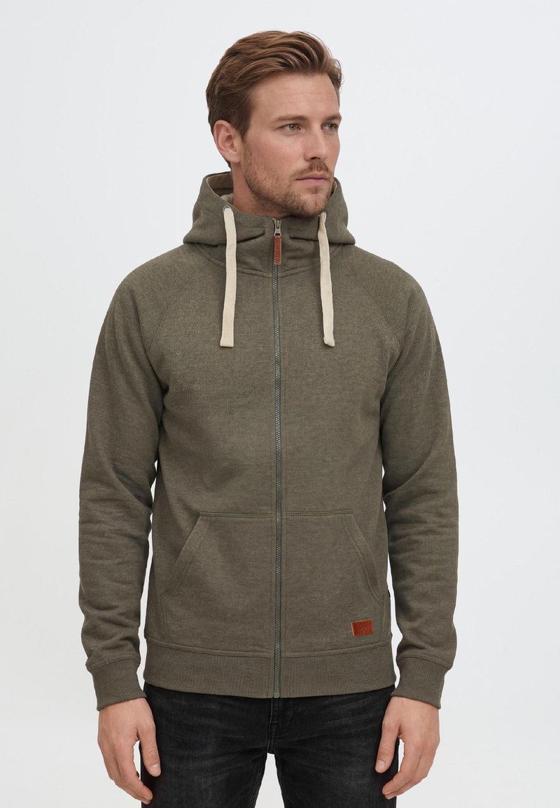 Man draagt een groene hoodie met rits en crèmekleurige trekkoorden, handen ontspannen langs het lichaam, kijkt iets naar rechts tegen een effen achtergrond.