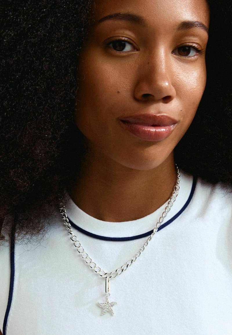 Collier en chaîne argentée avec un pendentif en forme d'étoile présentant des détails. Porté sur un haut blanc avec une bordure bleu marine. Texture lisse.