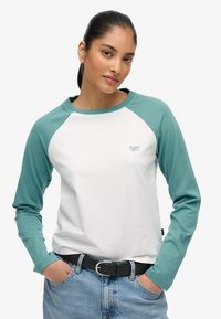 Wit shirt met raglanmouwen en turkooizen mouwen, gemaakt van katoen, met een klein groen logo op de borst, gecombineerd met blauwe denim.