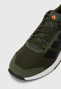 Vue de face d'une basket de sport verte en tissu mesh, lacets foncés, bandes de logo noires et semelle blanche sur un fond clair.