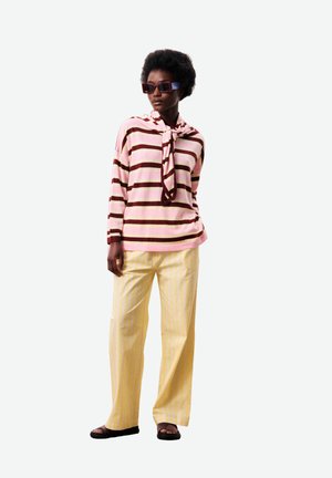 Vrouw die een roze en bruin gestreept shirt met lange mouwen draagt, gele wijd uitlopende broek, zwarte sandalen en donkere zonnebril, staand met ontspannen handen.