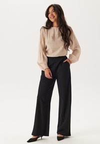 Beige textuur blouse met lange mouwen en een ronde hals, gecombineerd met hooggewrongen zwarte wijde broek. Zwarte hakken maken de outfit compleet.