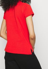 Polo shirt in cotone rosso con maniche corte, caratterizzato da spacco laterale all'orlo per maggiore libertà di movimento. Design semplice e classico, vista posteriore mostrata.