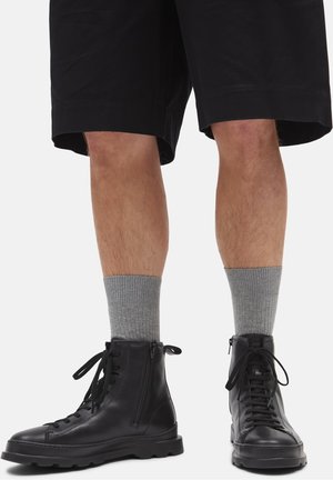 Benen in zwarte shorts, grijze geribbelde sokken en zwarte leren veterlaarzen met zijritsen op een witte achtergrond.