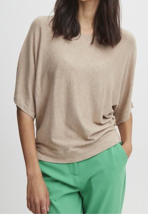 Femme portant un haut beige ample à manches courtes et un pantalon vert vif, une main dans la poche, sur un fond uni.