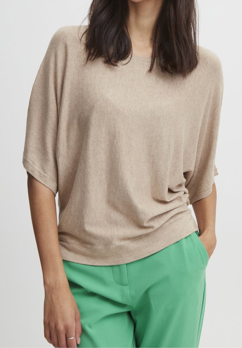 Femme portant un haut beige ample à manches courtes et un pantalon vert vif, une main dans la poche, sur un fond uni.