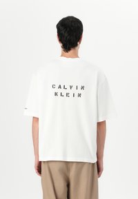 Balta medvilninė marškinėliai su trumpomis rankovėmis, su juodu blokiniu raštu "CALVIN KLEIN" ant nugaros ir minimaliais dizaino elementais.