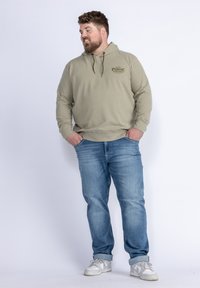 Felpa verde oliva chiaro con un logo sul petto, abbinata a jeans blu sbiaditi e sneakers bianche. Tessuto spesso, vestibilità comoda, cappuccio con coulisse.