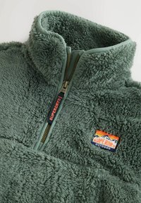 Grüner Fleece-Pullover mit hohem Kragen, Reißverschluss und strukturiertem Stoff. Mit einem bunten "Superdry Outdoor"-Logo-Patch.