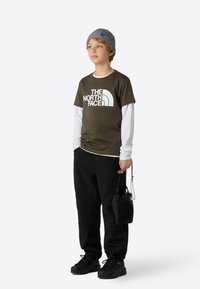 Olivovo zelené tričko s bielym logom, vrstvené cez dlhý biely rukáv. Čierne nohavice, čierne čižmy a sivá pletená čiapka vytvárajú kontrast.