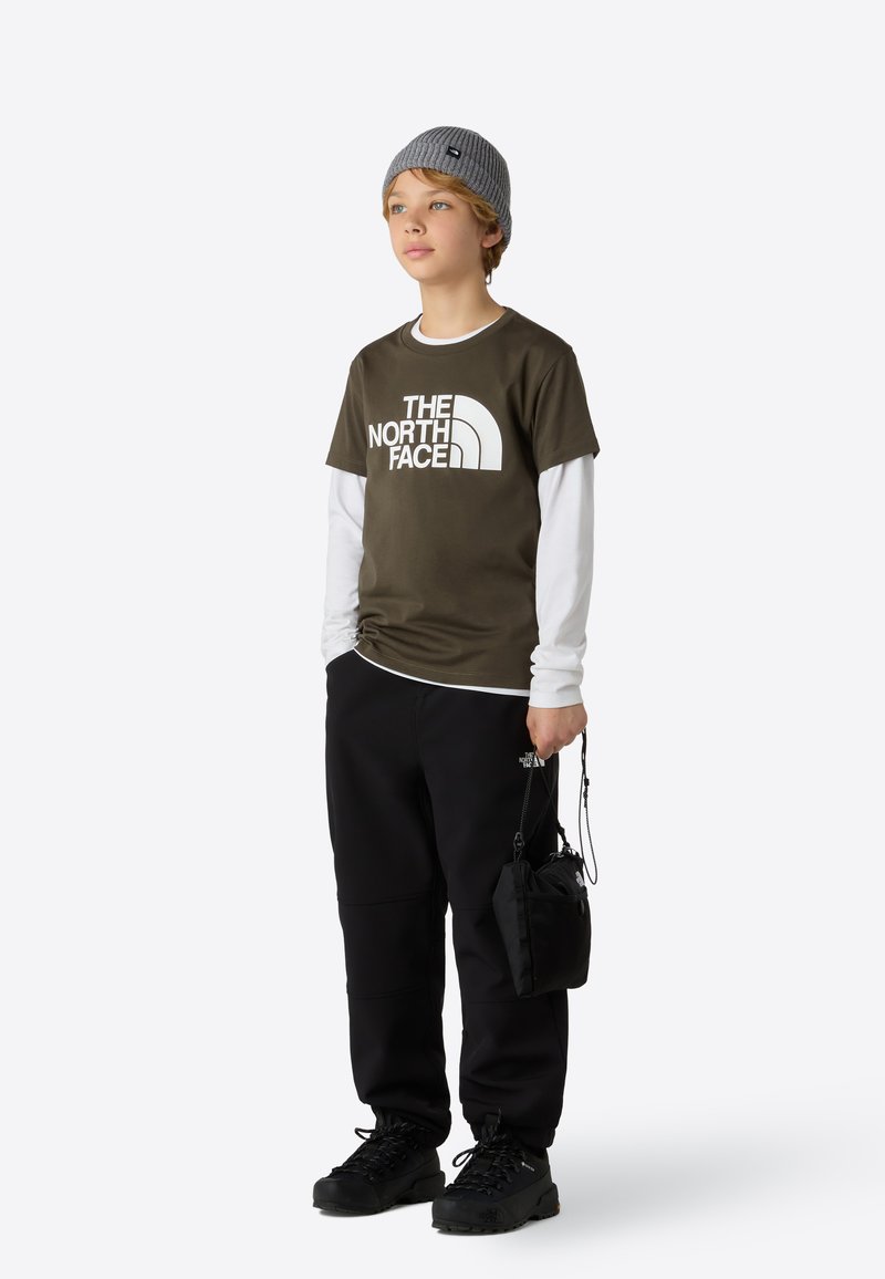 Olivovo zelené tričko s bielym logom, vrstvené cez dlhý biely rukáv. Čierne nohavice, čierne čižmy a sivá pletená čiapka vytvárajú kontrast.