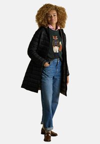 Žena s kučeravými vlasmi oblečená v čiernom prešívanom kabáte, svetre s grafiku líšky, ružovej košeli s golierom, modrých džínsoch a hnedých mokasínach s béžovými ponožkami.