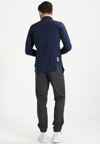 Man i en marinblå långärmad skjorta och mörkgrå joggingsbyxor med vita sneakers, stående med ryggen vänd mot kameran.