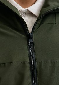 Giacca verde con una finitura testurizzata, caratterizzata da una zip nera e una camicia colletto chiara sotto.