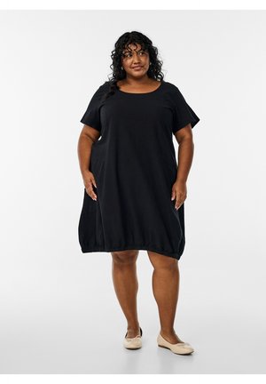 Sieviete ar cirtainiem matiem, tērpusies vaļīgā melnā kleitā ar īsām piedurknēm un valkājot bēša krāsas baleta kurpes, stāv pret vienkāršu fonu.