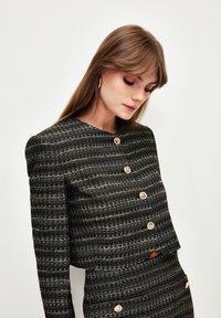 Chaqueta recortada a rayas negras y doradas con mangas largas, que presenta grandes botones redondos y una tela texturizada, creando una silueta estructurada.