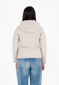 Giacca beige con cappuccio, dal design aderente, texture morbida e polsini elasticizzati, abbinata a jeans di denim a vita alta di colore blu chiaro.