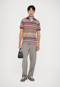 Polo a righe multicolore con colletto bordeaux, abbinato a pantaloni grigi chiari. Il modello tiene una borsa nera e indossa scarpe da ginnastica verdi.