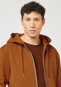 Giovane uomo con capelli ricci e scuri che indossa una felpa marrone con zip sopra una maglietta marrone, rivolto in avanti con un'espressione neutra.