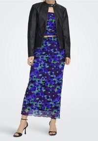 Giubbotto di pelle nera sopra un vestito maxi con motivo floreale viola e verde, con corpetto aderente e gonna fluida, abbinato a tacchi neri a strappo.