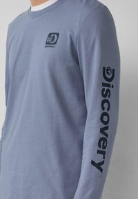 Lange mouwen, lichtblauw katoenen T-shirt met een klein logo aan de voorkant en grote 'Discovery' tekst in zwart gedrukt langs de linker mouw.