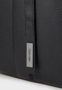 Čierna kožená taška s textúrovaným povrchom, s striebornou obdĺžnikovou štítkou s vyrytým názvom "Calvin Klein" a švom.