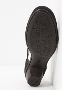 Chaussure noire avec une semelle en caoutchouc texturée, un petit talon bloc et un design ouvert comprenant une découpe latérale et des coutures décoratives.