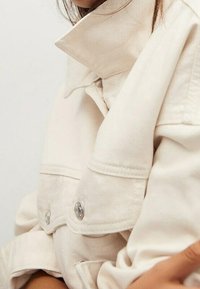 Beige denim jacket with a high collar and two front pockets, featuring metallic buttons and subtle stitching details. Smooth texture.