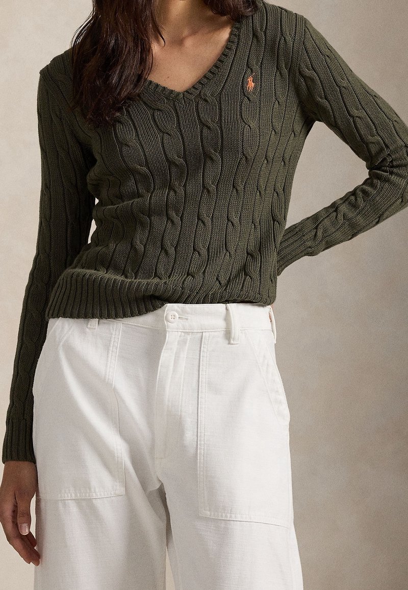 Personne portant un pull vert foncé à col en V en maille torsadée avec un pantalon blanc taille haute à jambes larges, debout avec une main sur la hanche.