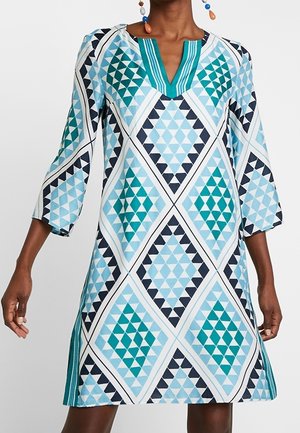 Frau trägt ein dreiviertelärmliges Kleid mit einem geometrischen dreieckigen Muster in Blau, Türkis und Schwarz sowie einem gestreiften V-Ausschnitt.